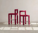 Two modern bar stools with thick, blocky magenta-pink frames and matching upholstered seats, standing on a concrete floor against a textured plaster wall with soft window shadows.