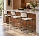 Three modern bar stools with cream textured upholstery and chrome cantilever legs lined up at a kitchen island with a marble countertop and wood cabinetry.