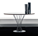 An oval dining table with a dark wood top and a sculptural wire pedestal base, shown on a black rug in front of a white wall. Two dark decorative vases sit on the tabletop.