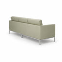 A modern three-seat sofa in a light beige leather or faux leather upholstery with a slim chrome metal frame and legs, shown from a three-quarters perspective against a plain white background.