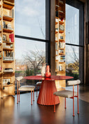 Midj Plissé 59-inch round dining table with red pleated pedestal base beside tall windows and bookshelves