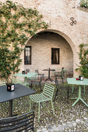 Midj Trampoliere outdoor slatted metal side chairs in charcoal and sage green at cafe tables in a brick courtyard